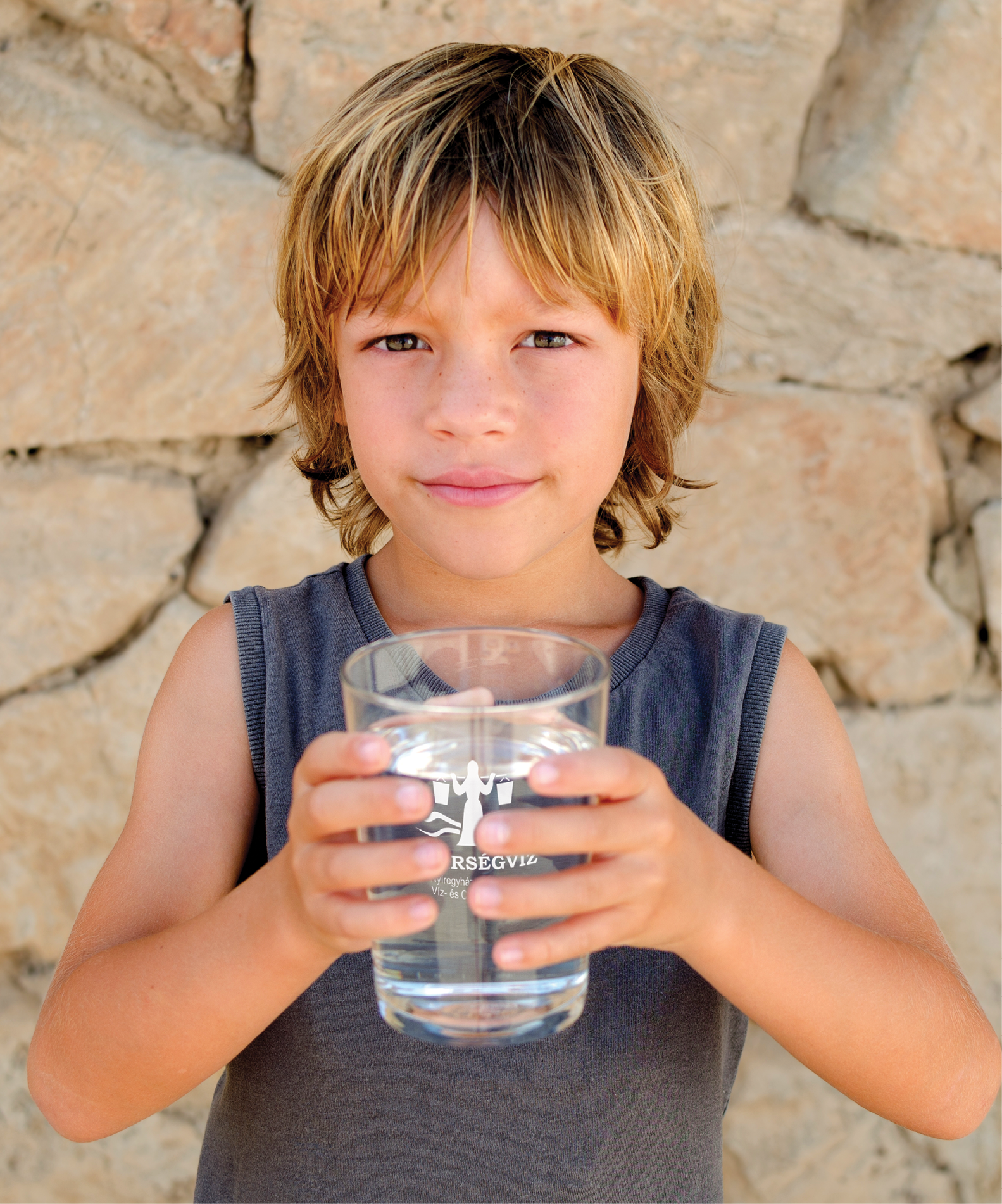 Horizontal photography of adorable blond caucasian child looking at camera holding a glass of pure mineral water outdoors in summer. Healthy lifestyle and refreshment concept. Copy space